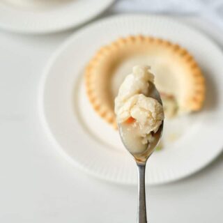 An aerial image of a golden brown pot pie sitting on a white dinner plate with a bite out of it. A woman's hand is holding a spoon filled with the pot pie contents over the cooked pie below.