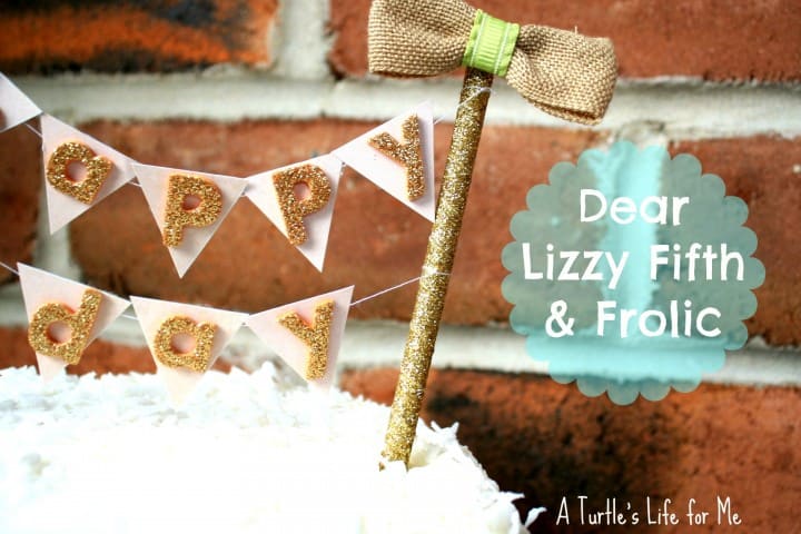 A homemade cake bunting with pennant flags made from the Dear Lizzy Fifth and Frolic craft line. The bunting white and gold bunting sits on a white frosted cake against a brick wall. There is text overlaid that says Dear Lizzy Fifth and Frolic Cake Bunting. 