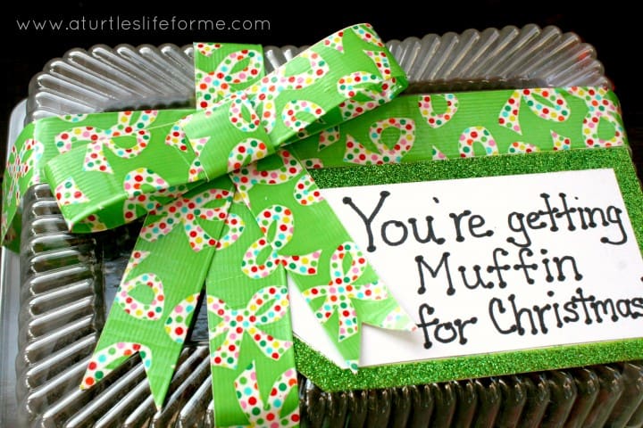 A close cropped photo of a closed disposable cupcake carrier that is decorated with green ribbon and a hand written label that says You're Getting Muffin for Christmas. 