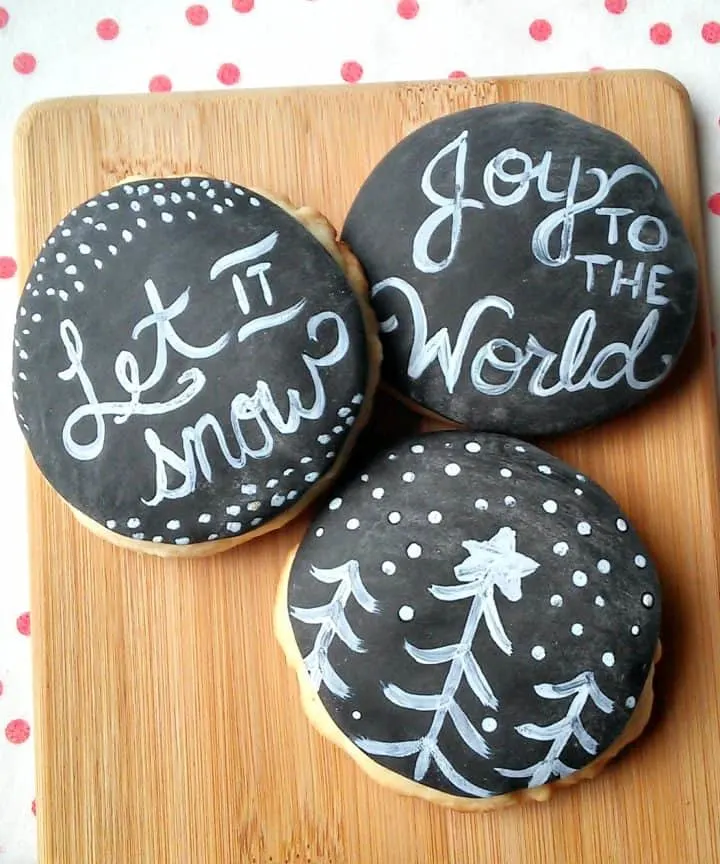 Three sugar cookies are displayed on a wooden cutting board.  The cookies are covered in black fondant and decorated to look like chalkboards, with holiday messages and decorations for Christmas.