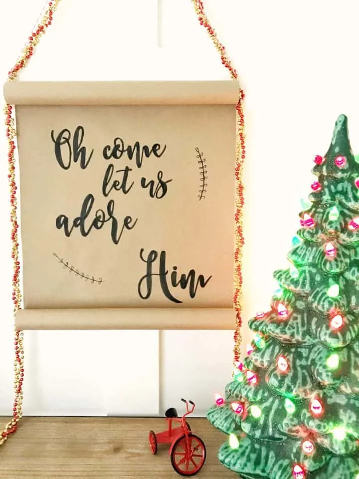 A hand lettered Christmas scroll made from kraft paper hangs against a white wall next to  a vintage  Christmas ceramic tree. The text on the scroll says, "Oh Come Let Us Adore Him"