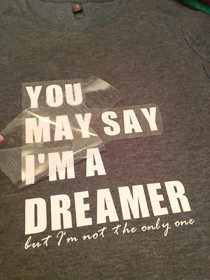 How to make a band tshirt with vinyl. A process shot showing the lyrics You May Say I'm a Dreamer but I'm Not the Only One made into a Cricut vinyl transfer with white text before being ironed onto a grey tshirt
