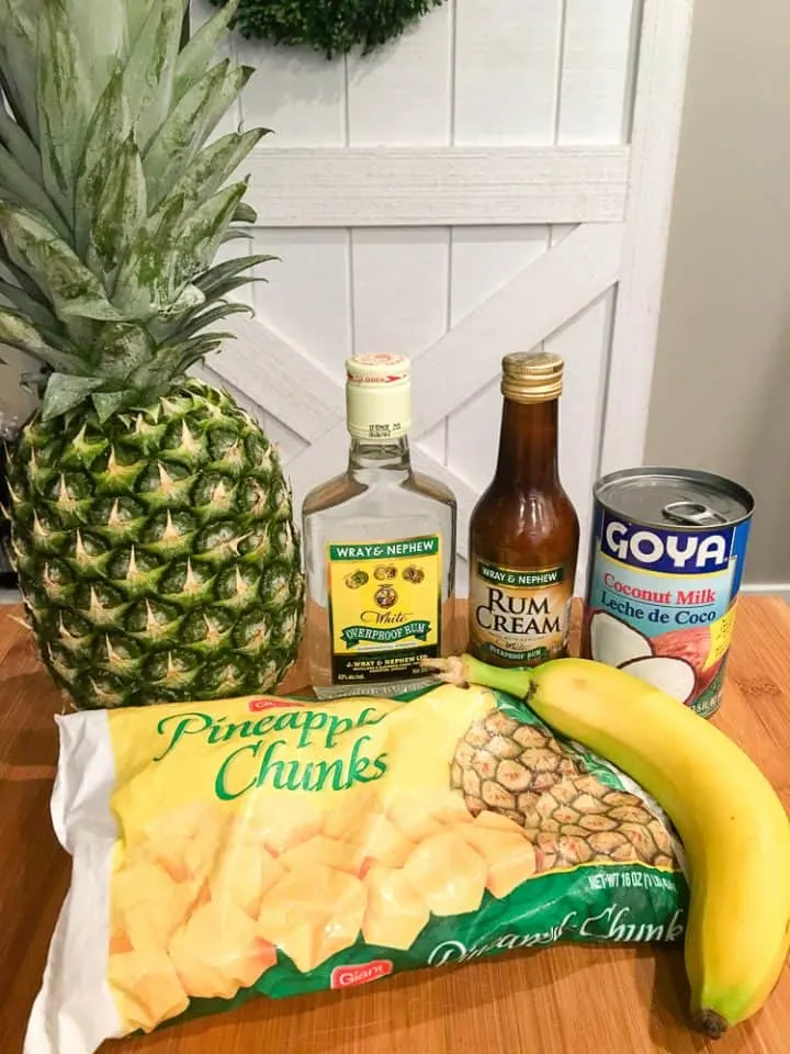 Dole Whip with Rum Recipe Ingredients on a table, including rum, rum cream, coconut milk, a fresh pineapple, frozen pineapple chunks, and a banana