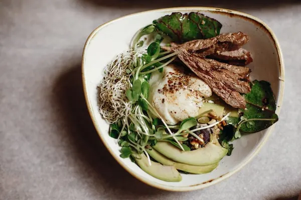 Delicious salad with juicy grill steak, avocado, sprouted greens, leaves, egg pouch, nuts in stylish bowl on a table.