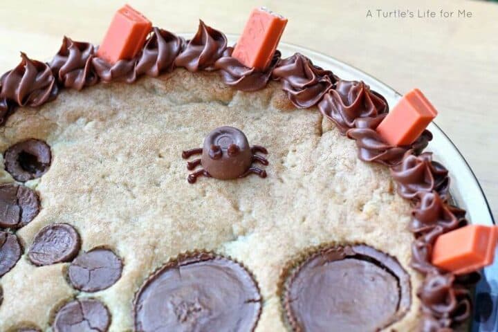 A detailed photograph of a spider made from a Rolo chocolate candy and frosting sitting atop a cookie pie that is Halloween themed