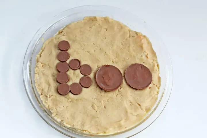 A process shot of a cookie pie being made, showing the cookie pressed into a baking dish and the word "boo" spelled out with rolo candies and 2 chocolate peanut butter cups