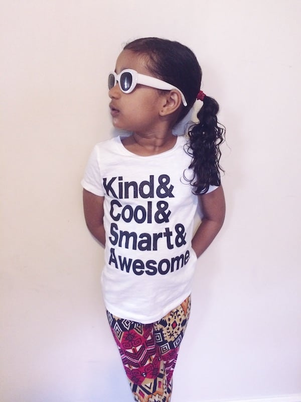 A young girl wearing white sunglasses and a white tshirt with the text  Kind & Cool & Smart & Awesome. She is  looking to the side and standing against a white wall. 