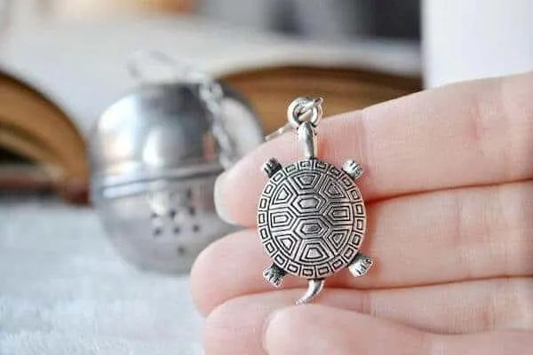 A woman's hand  holds a silver turtle charm attached to a tea ball, which is visible in the background