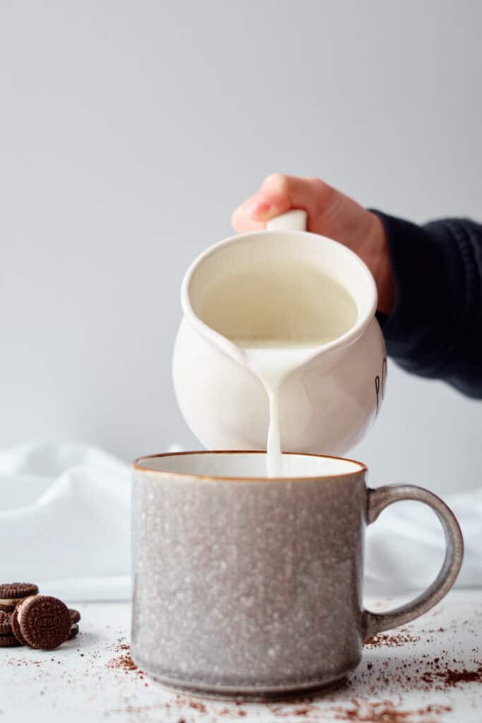 A hand is seen pouring hot milk from a white milk jug into a mug. The mug sits on a white table with mini Oreos and hot chocolate powder strewn on the table. 