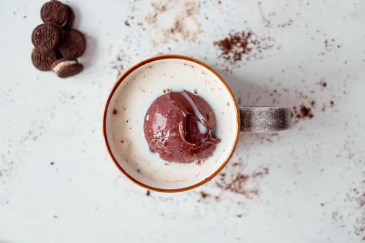 A hot chocolate sphere  begins to melt in a mug of hot milk. The photo is birds eye view, and the mug sits on a white table with hot chocolate mix and mini Oreos strewn on the table too