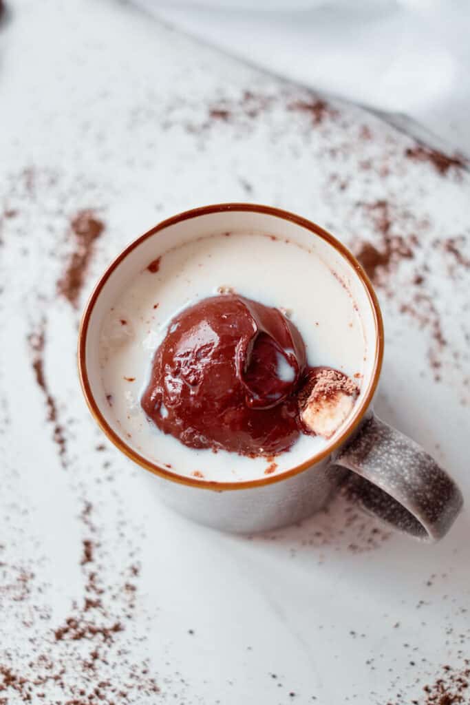 A homemade hot cocoa bomb melts in a mug of hot milk. A mini marshmallow can be seen erupting from the hot chocolate bomb.