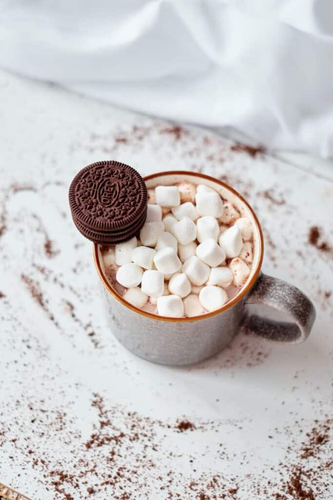 A mug of hot chocolate made using a hot cocoa bomb. Pictured is a mug of hot chocolate topped with mini marshmallows and a stack of mini Oreos against an isolated white background.