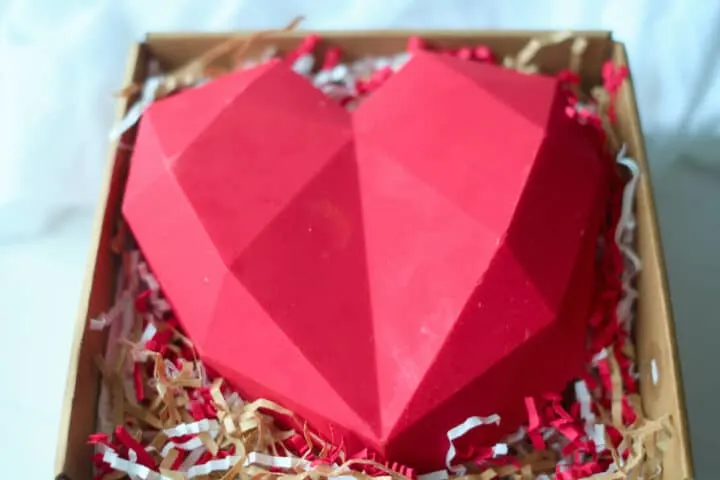 An edible chocolate box made from red candy melts sits in a box on a bed of shredded paper