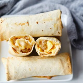 A cooked breakfast burrito that has been cut in half. The two halves are sitting side by side on a plate with other uncut burritos, and the viewer can see cooked eggs, breakfast sausage crumbles and melted cheese within the burrito.