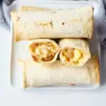 A cooked breakfast burrito that has been cut in half. The two halves are sitting side by side on a plate with other uncut burritos, and the viewer can see cooked eggs, breakfast sausage crumbles and melted cheese within the burrito.