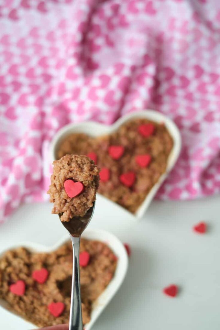 A spoon filled with a mouthful of chocolate oatmeal and a handmade melting wafer heart in the foreground. Chocolate oatmeal in a heart shaped bowl and garnished with red candy hearts in the background.