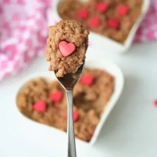 A spoon filled with a mouthful of chocolate oatmeal and a handmade melting wafer heart in the foreground. Chocolate oatmeal in a heart shaped bowl and garnished with red candy hearts in the background.