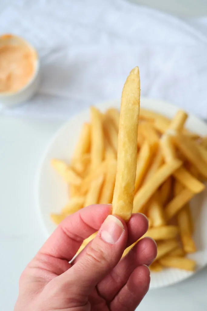 A woman's hand holds a french fry up with a plate of fries in the background