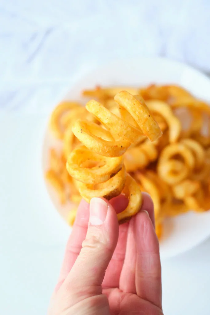 A woman's hand holds up a frozen curly fry cooked in air fryer with a plate of curly fries in the background