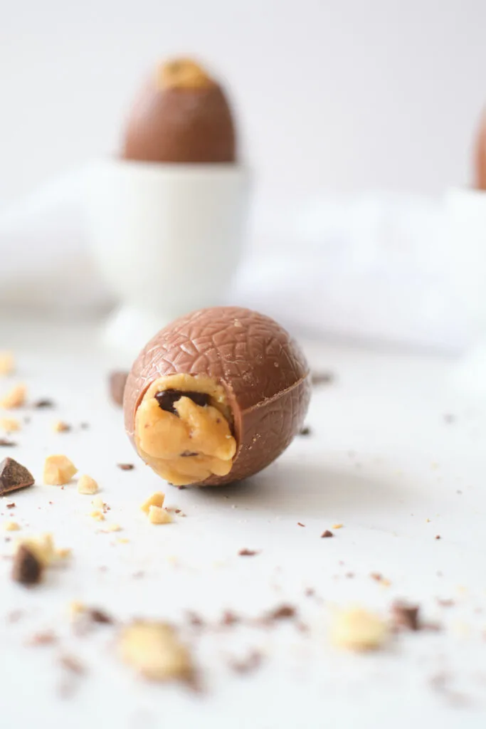 A peanut butter and chocolate chunk filled easter egg lies on its side on a white counter, with peanuts and chocolate chunks strewn around it