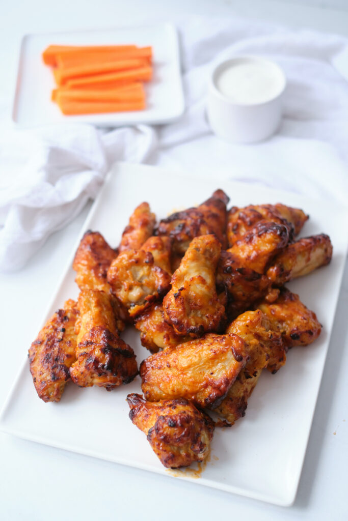 A white dinner plate piled high with buffalo chicken wings, with fresh carrot sticks and dip in the background. The photo shows an after shot for a tutorial about how to cook frozen chicken wings in air fryer.