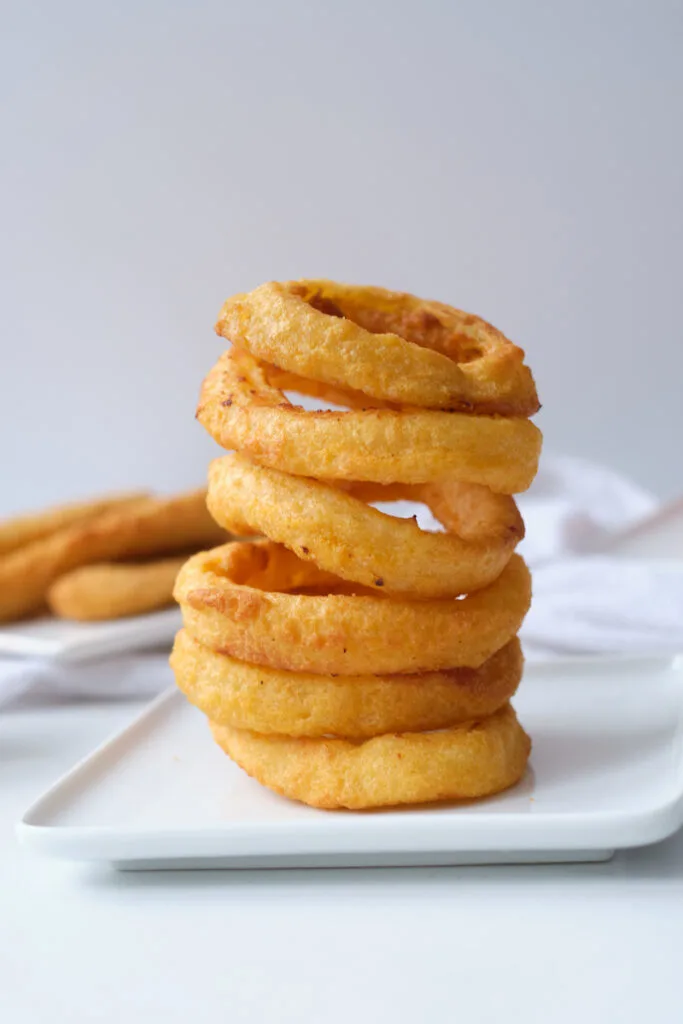 Stack of air fryer onion rings on a white dinner plate with chicken tenders in the background and out of focus