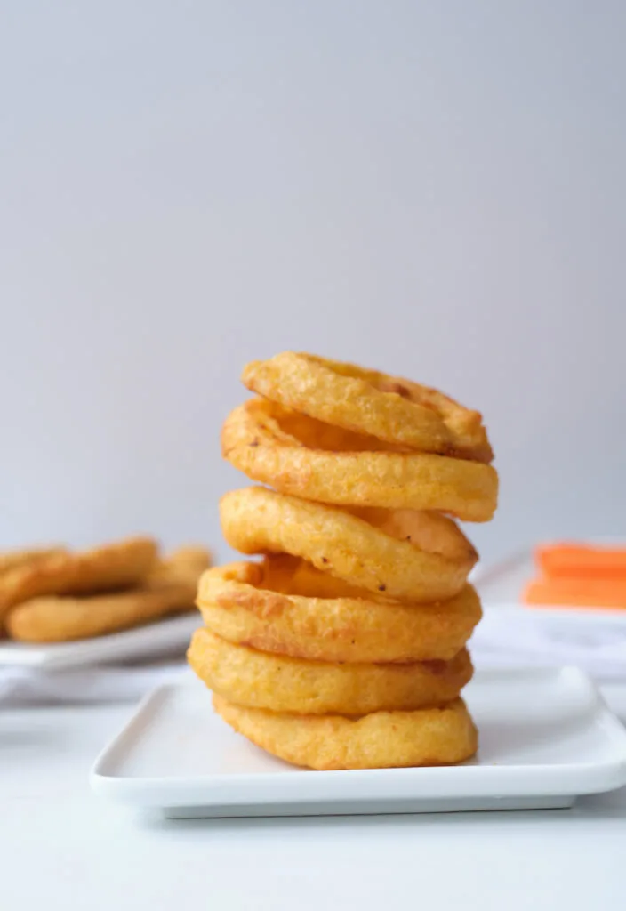 Stack of air fryer onion rings on a white dinner plate, with chicken tenders and carrot sticks in the background out of focus