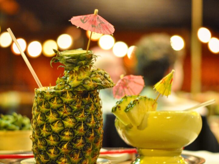 A cocktail in a pineapple and a pineapple cocktail in a glass with tropical umbrellas, with blurred background of a restaurant or lounge visible behind